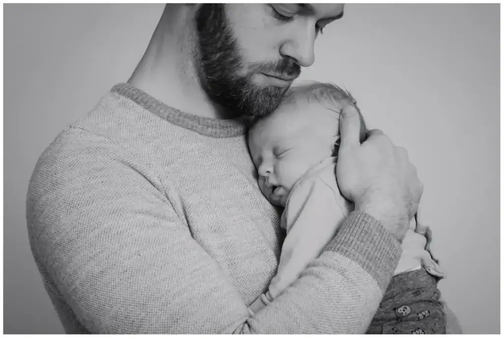 Père serrant son nouveau-né endormi contre lui pendant une séance photo de naissance en noir et blanc.