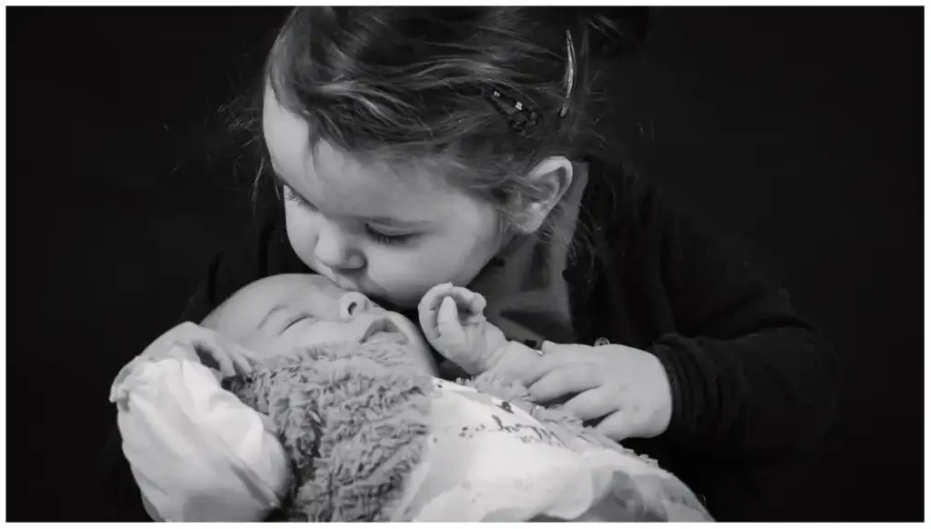 Jeune fille embrassant un nouveau-né dans ses bras lors d'une séance photo de naissance en noir et blanc.