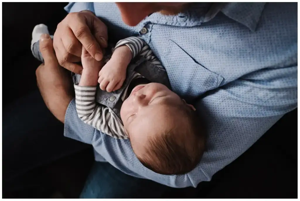 Père tenant son nouveau-né endormi dans ses bras pendant une séance photo de naissance sur fond sombre.
