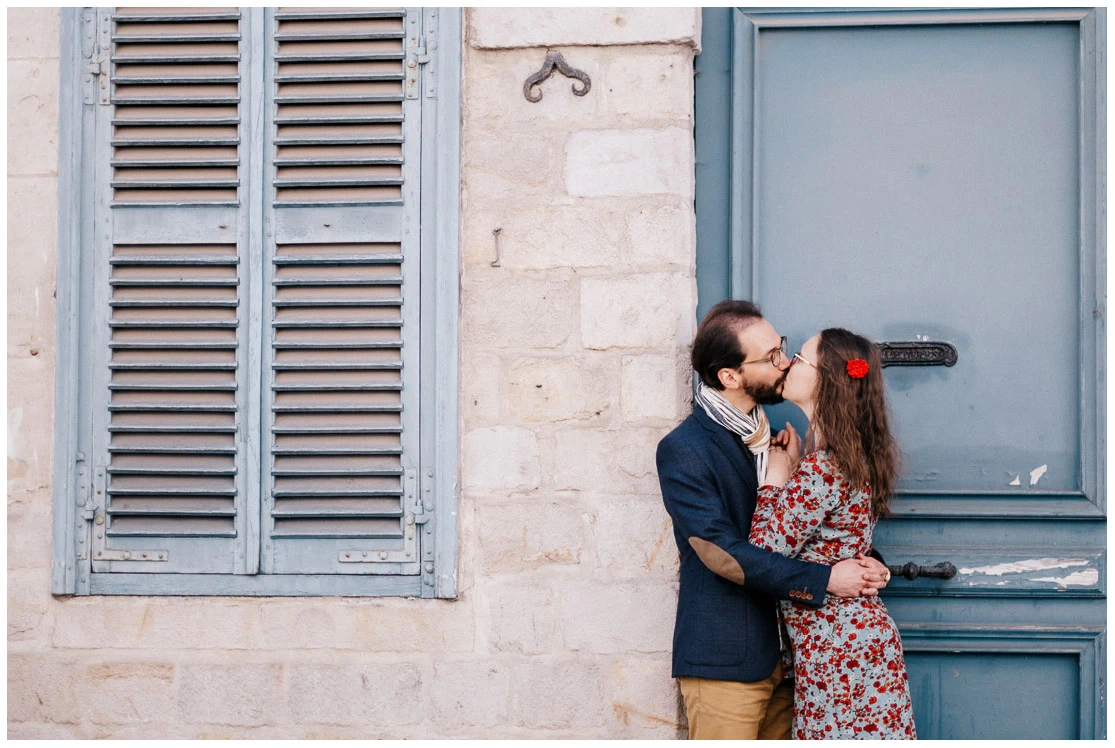 Couple s’embrassant devant une façade claire aux menuiseries bleu gris lors d’une séance photo lifestyle.