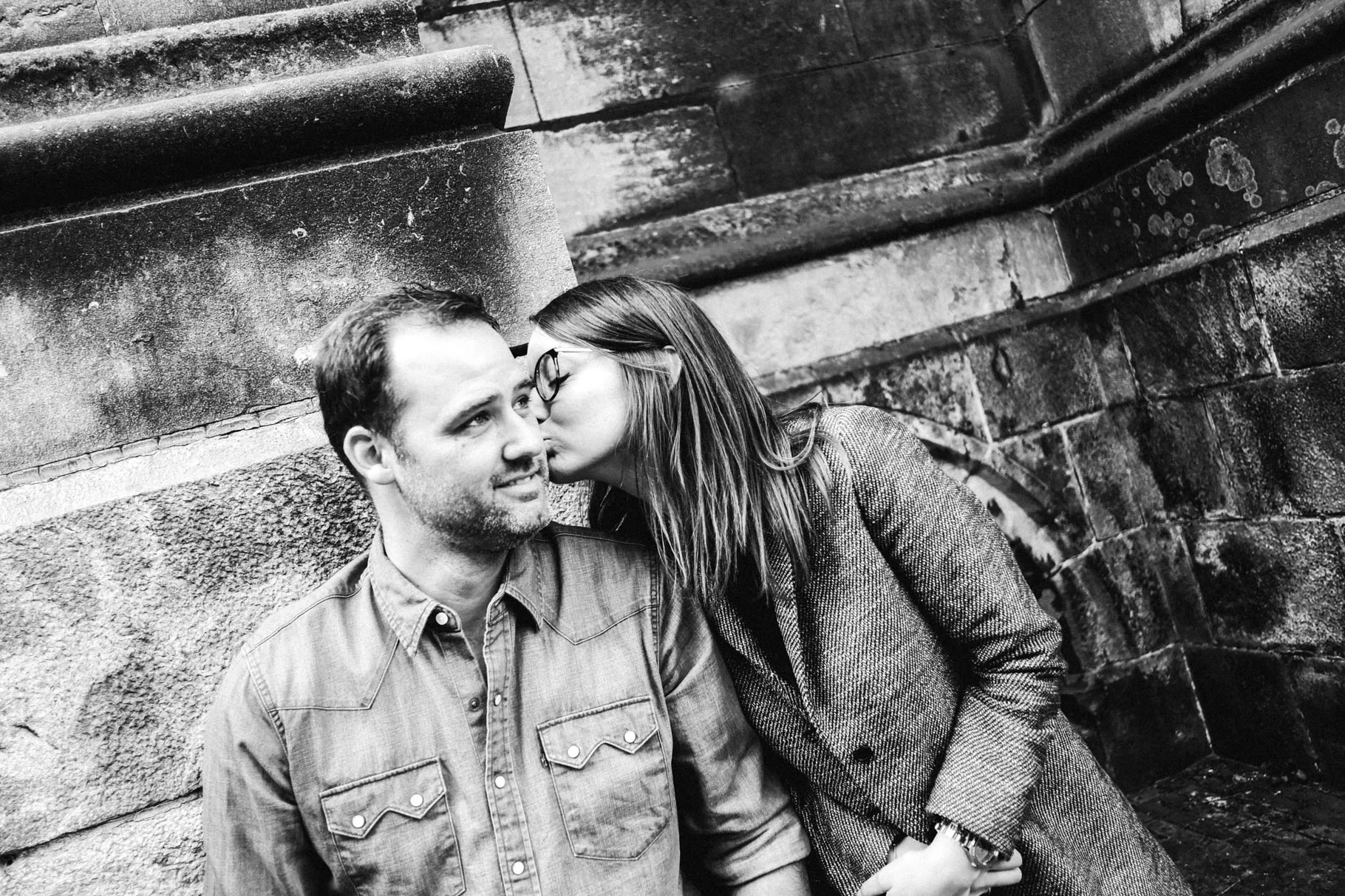 Femme se penchant tendrement vers son compagnon devant un mur en pierre, en noir et blanc.