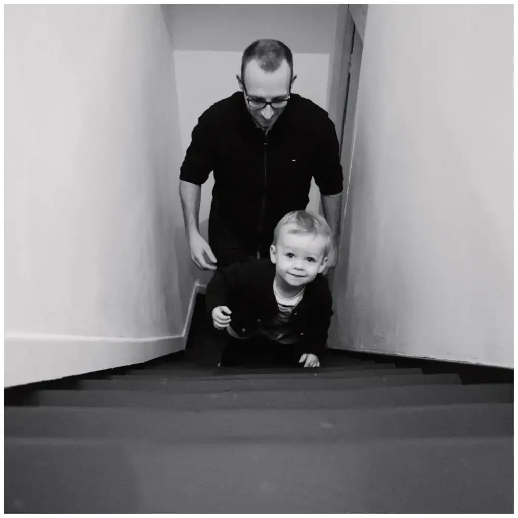 Un père accompagne son jeune enfant dans un escalier, dans une photographie en noir et blanc.