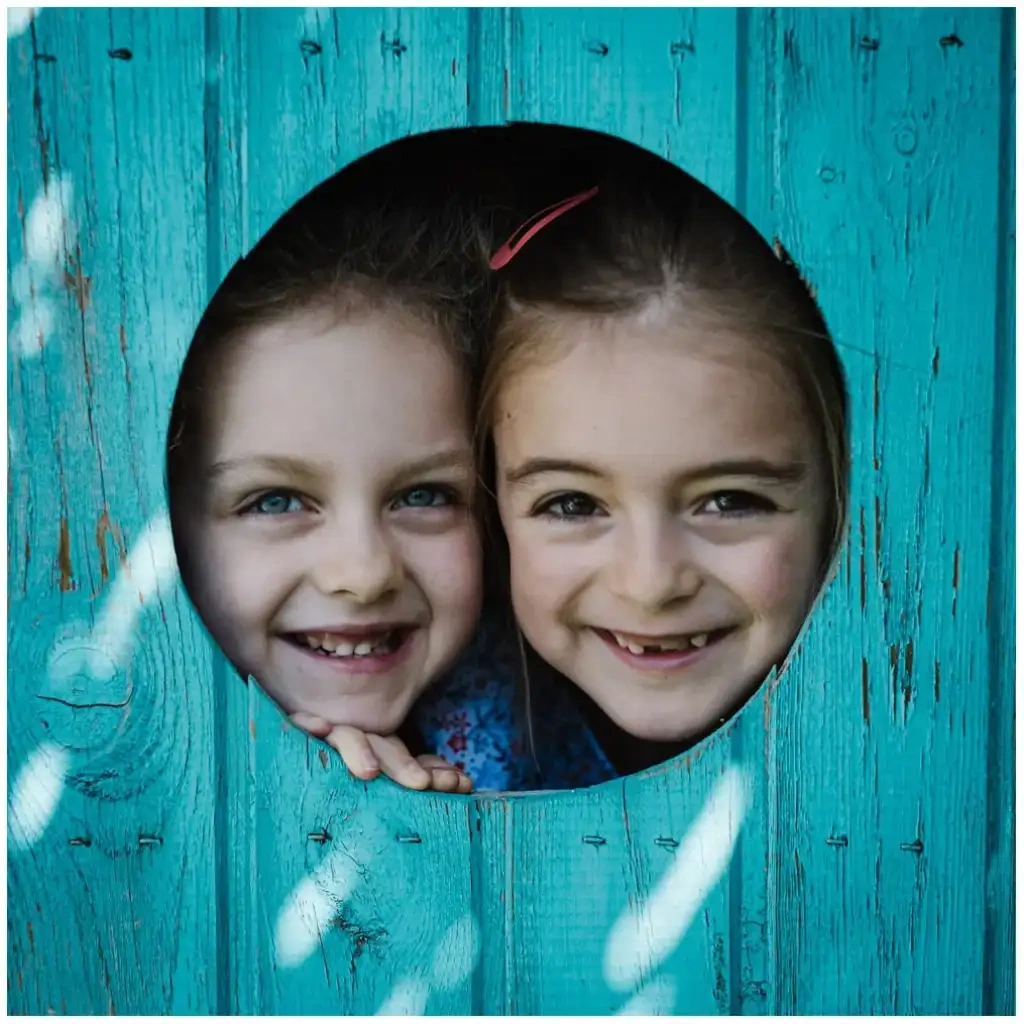 Deux jeunes filles souriantes regardent à travers une ouverture ronde dans une paroi turquoise.