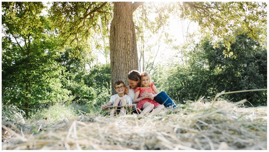 Mère entourant ses deux enfants assis dans l'herbe au pied d'un arbre.