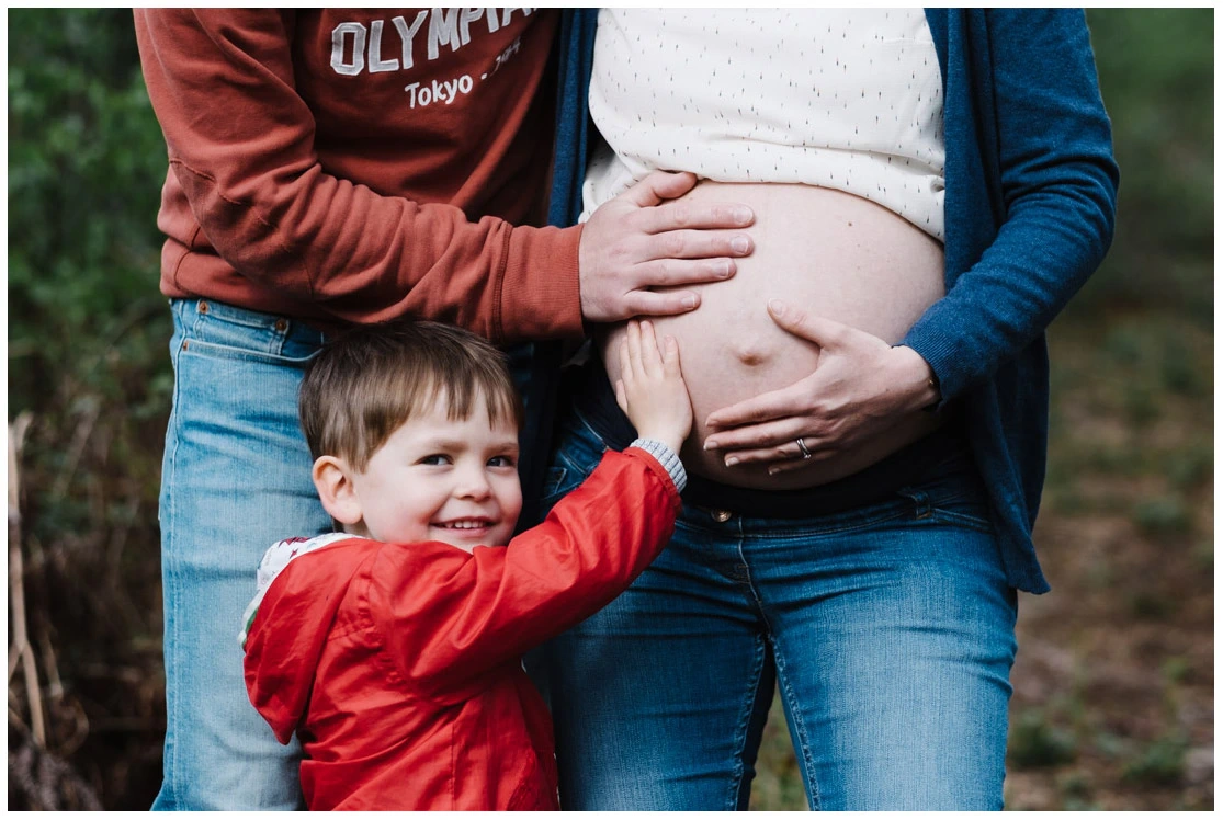 Jeune garçon posant sa main sur le ventre de sa mère enceinte pendant une séance photo de grossesse en famille.
