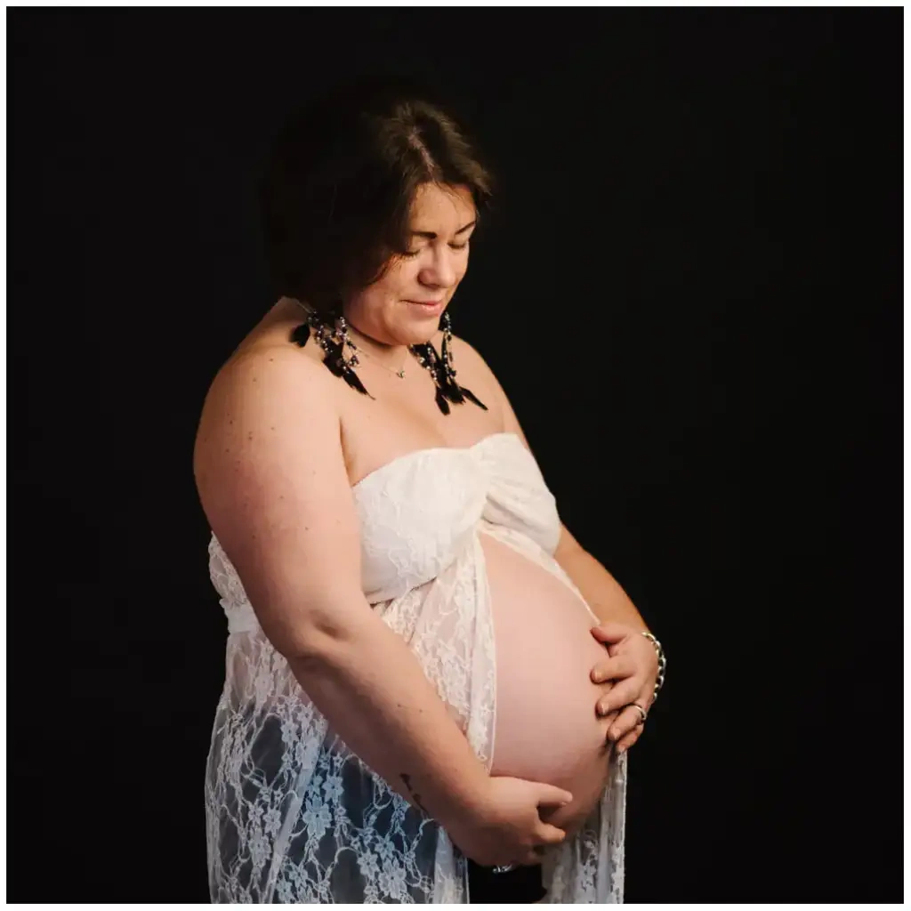 Une femme enceinte vêtue de dentelle blanche regarde son ventre devant un fond noir.