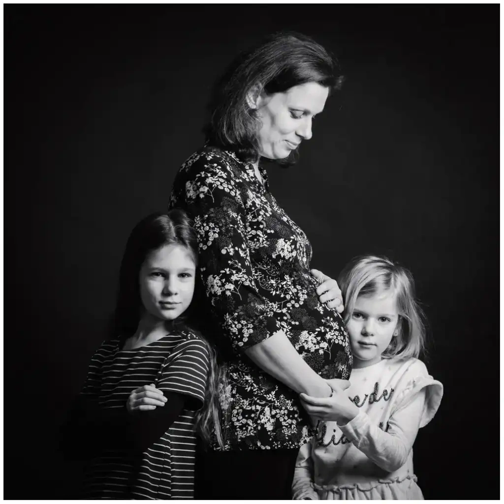 Portrait en noir et blanc d’une femme enceinte entourée de ses deux filles devant un fond sombre.