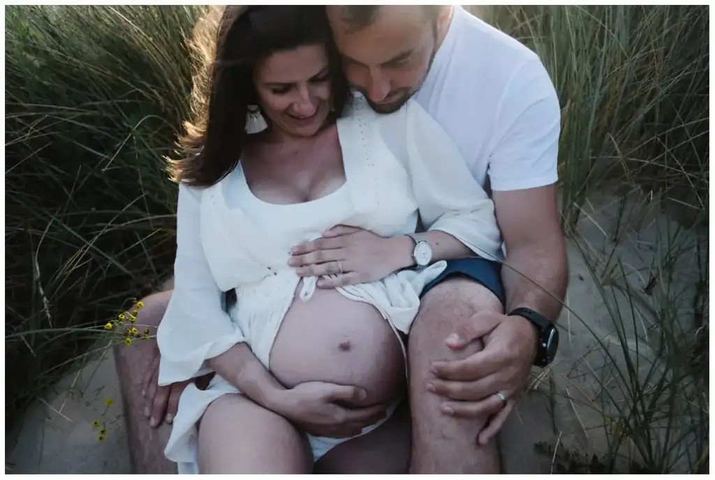 Un couple assis dans les dunes entoure le ventre de grossesse de ses mains.