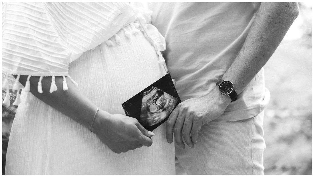 Couple tenant une échographie devant le ventre de la future mère pendant une séance photo de grossesse en noir et blanc.