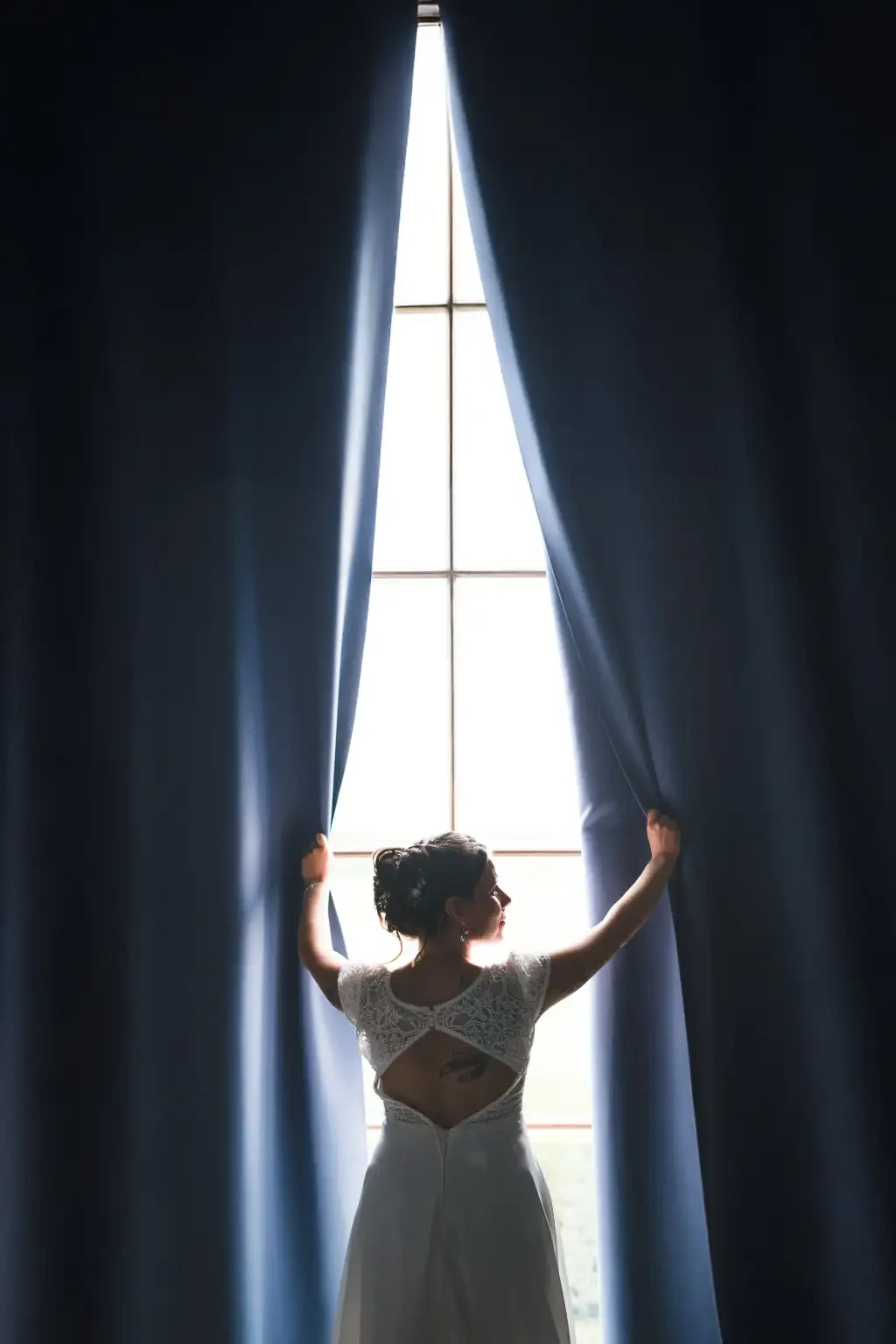 Mariée vue de dos ouvrant des rideaux bleu nuit devant une haute fenêtre lumineuse.