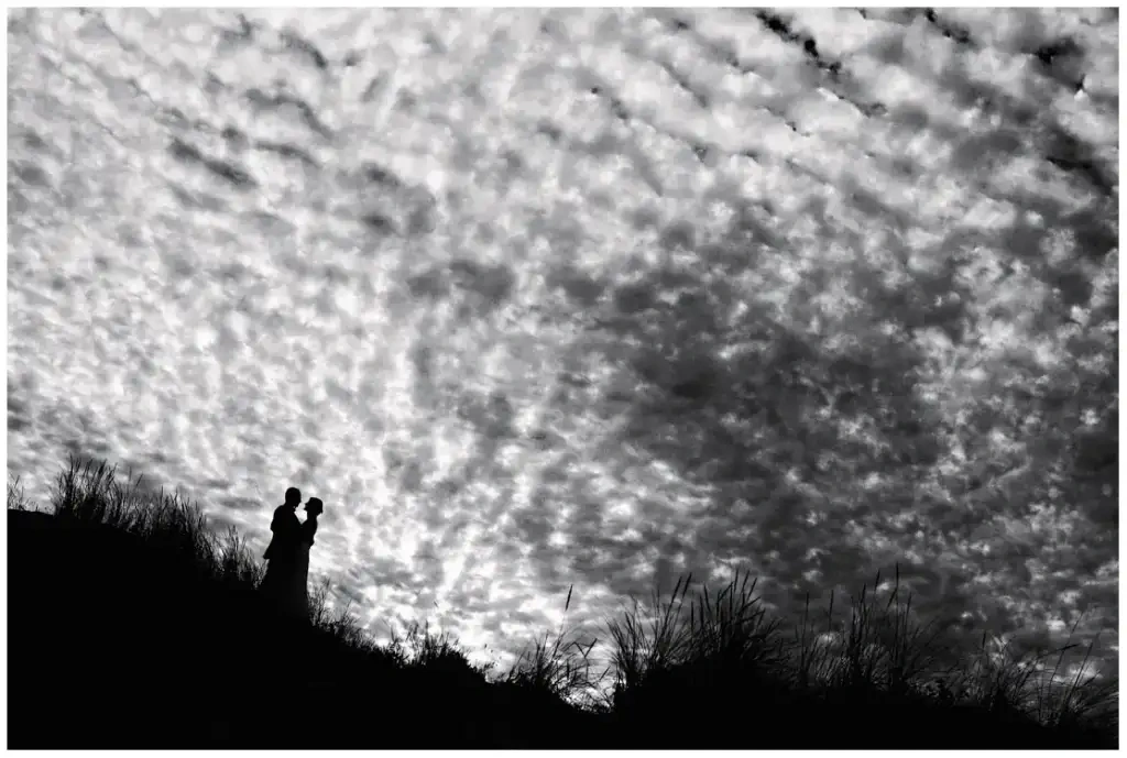 Silhouette en noir et blanc de mariés face à face sous un vaste ciel nuageux.