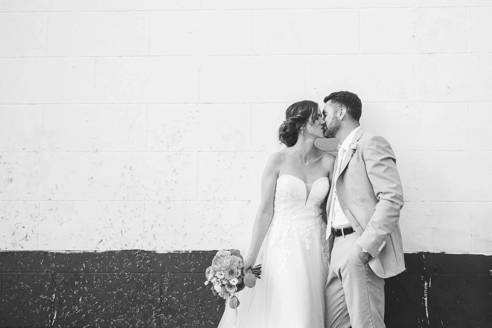 Mariés s'embrassant devant un mur clair, la mariée tenant son bouquet, lors d'un reportage de mariage.
