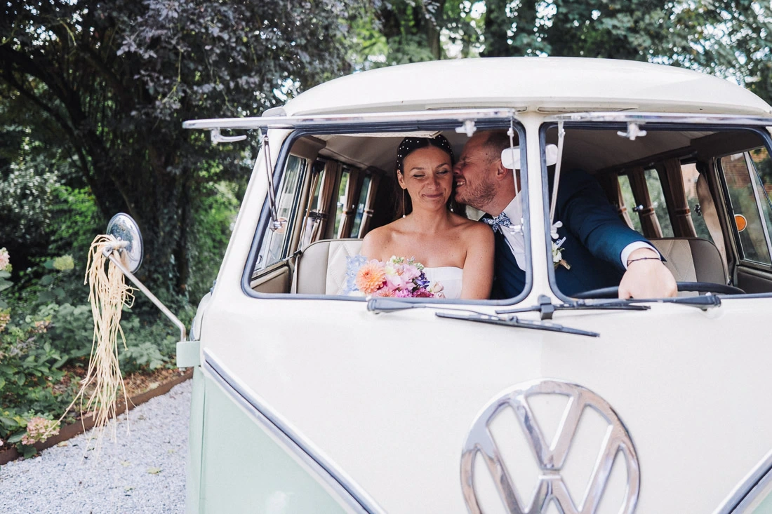 Mariée tenant son bouquet tandis que le marié l'embrasse dans un combi Volkswagen ancien.