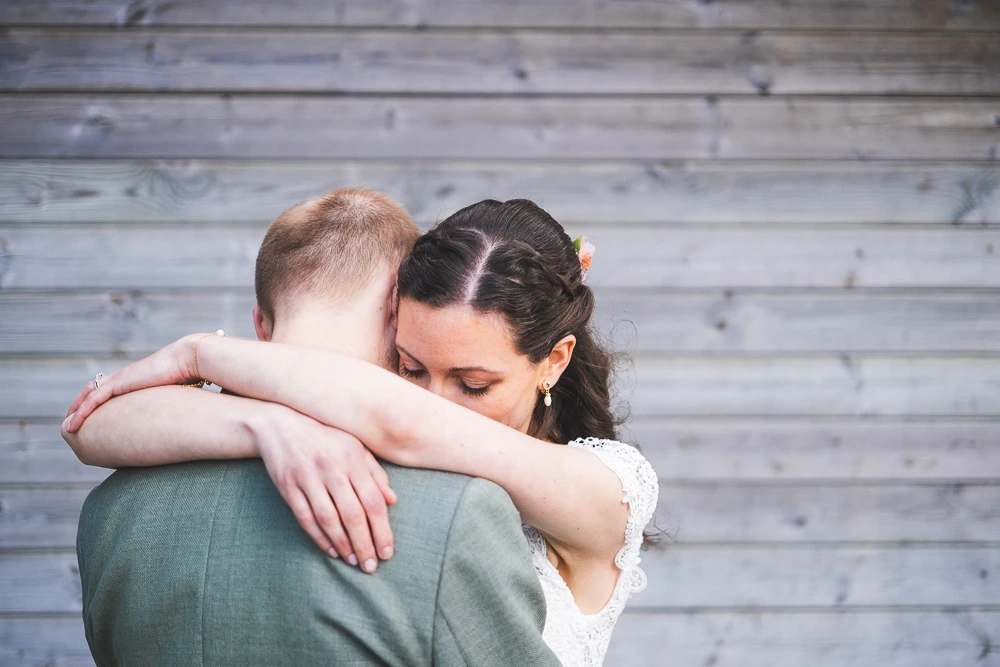Mariée enlacée avec le marié devant un mur en bois gris lors d'un reportage de mariage.