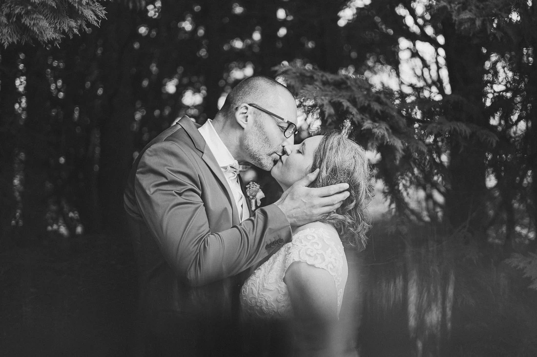 Mariés s'embrassant sous les arbres dans une photographie de mariage en noir et blanc.