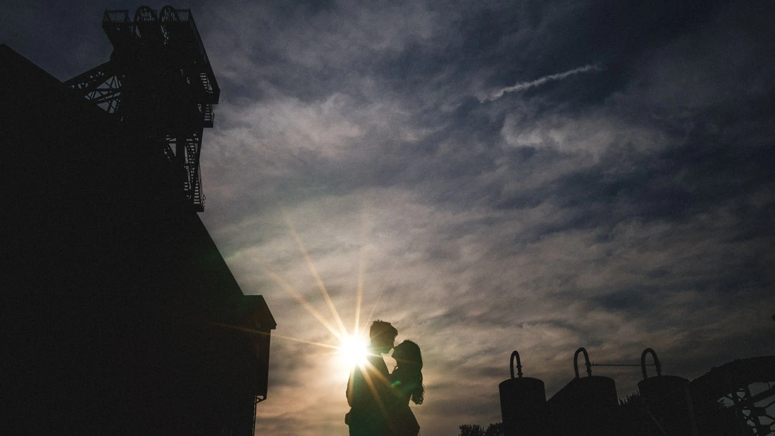 Silhouette de mariés s'embrassant devant le soleil sous un ciel nuageux.