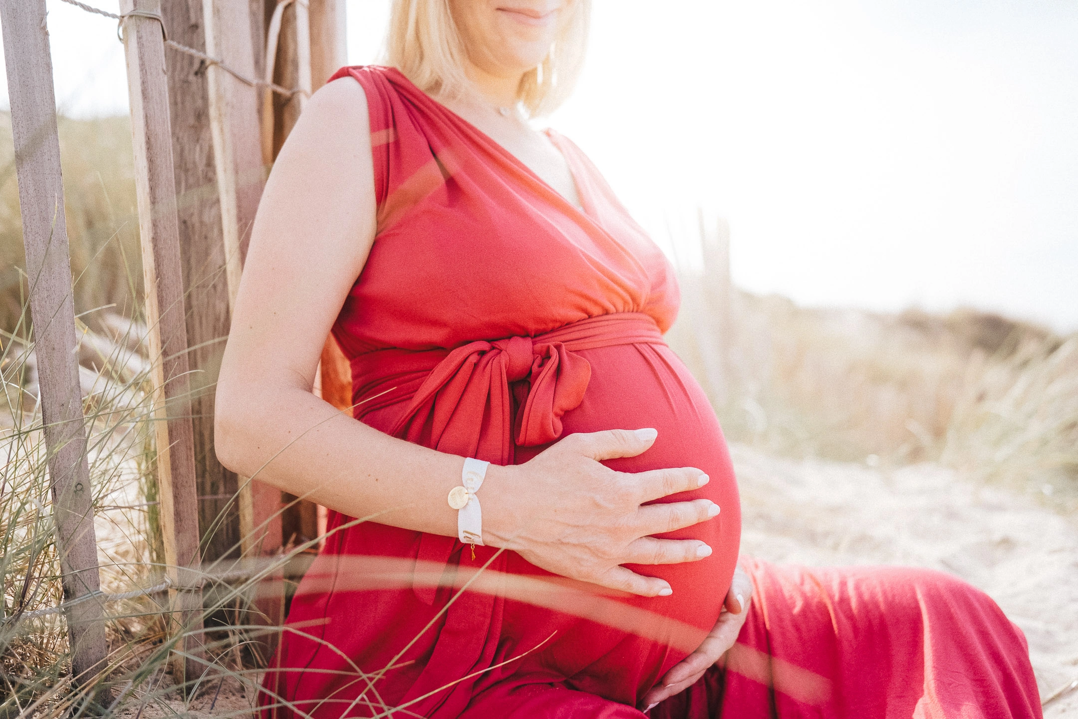 Femme enceinte en robe rouge assise dans les dunes, les mains posées sur son ventre.