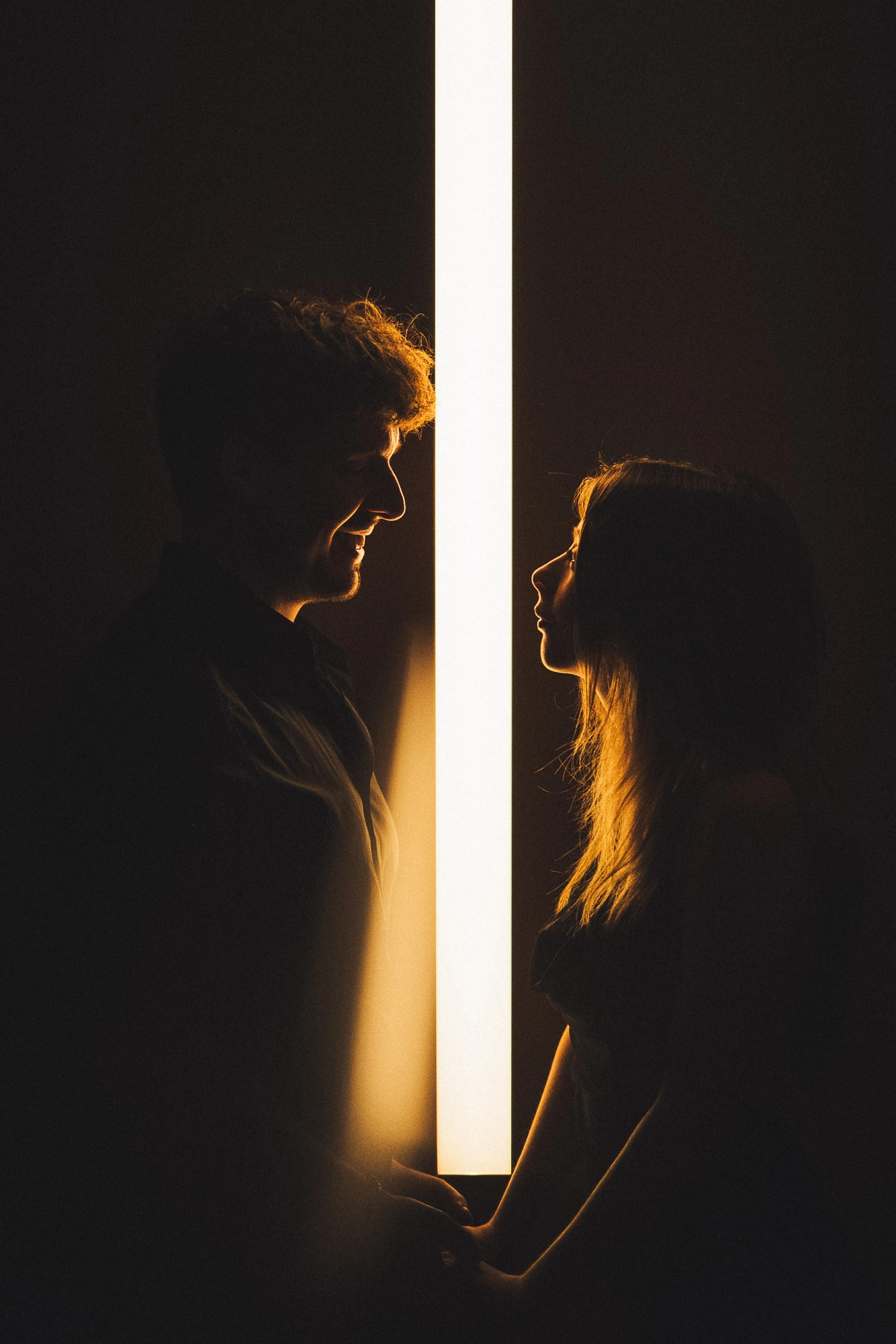 Couple se regardant de part et d’autre d’une barre lumineuse verticale sur fond sombre.