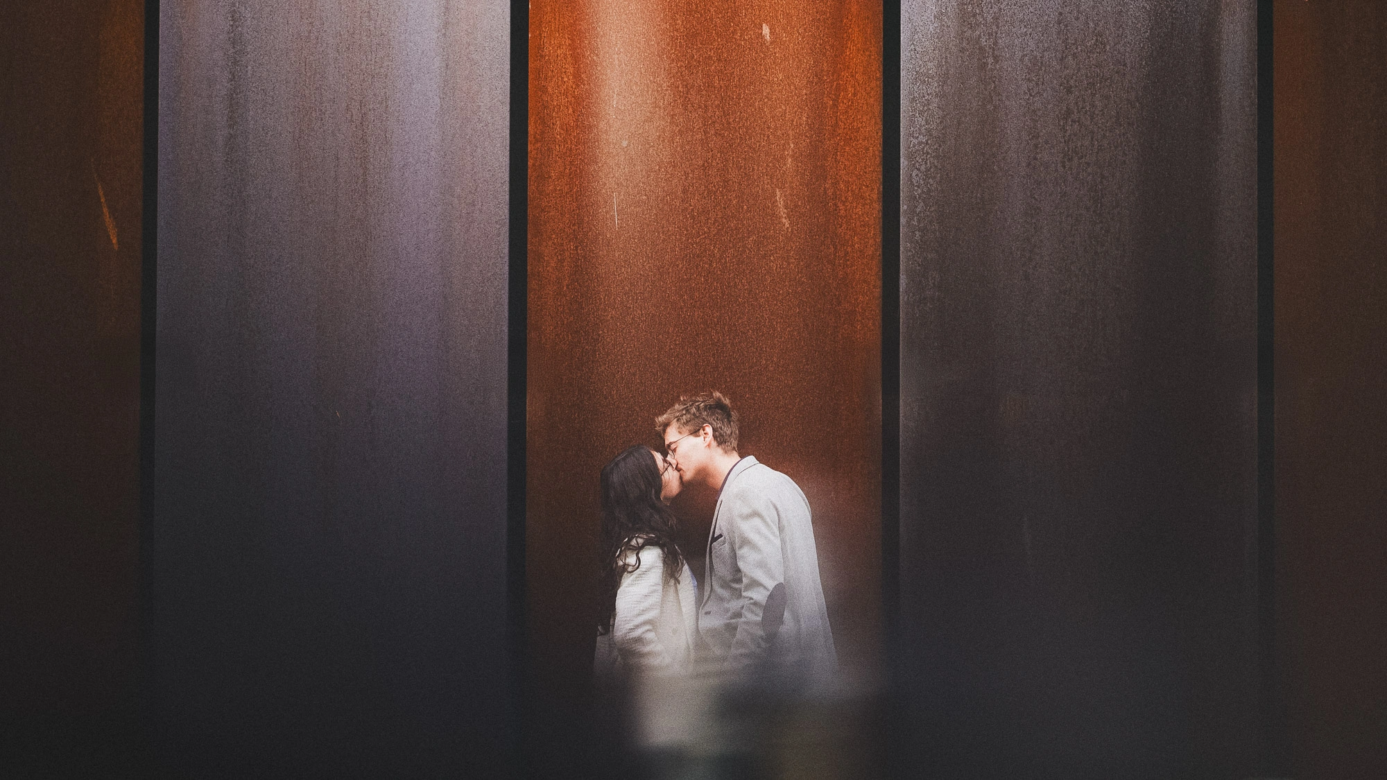 Couple s’embrassant devant de hauts panneaux architecturaux gris et rouille.