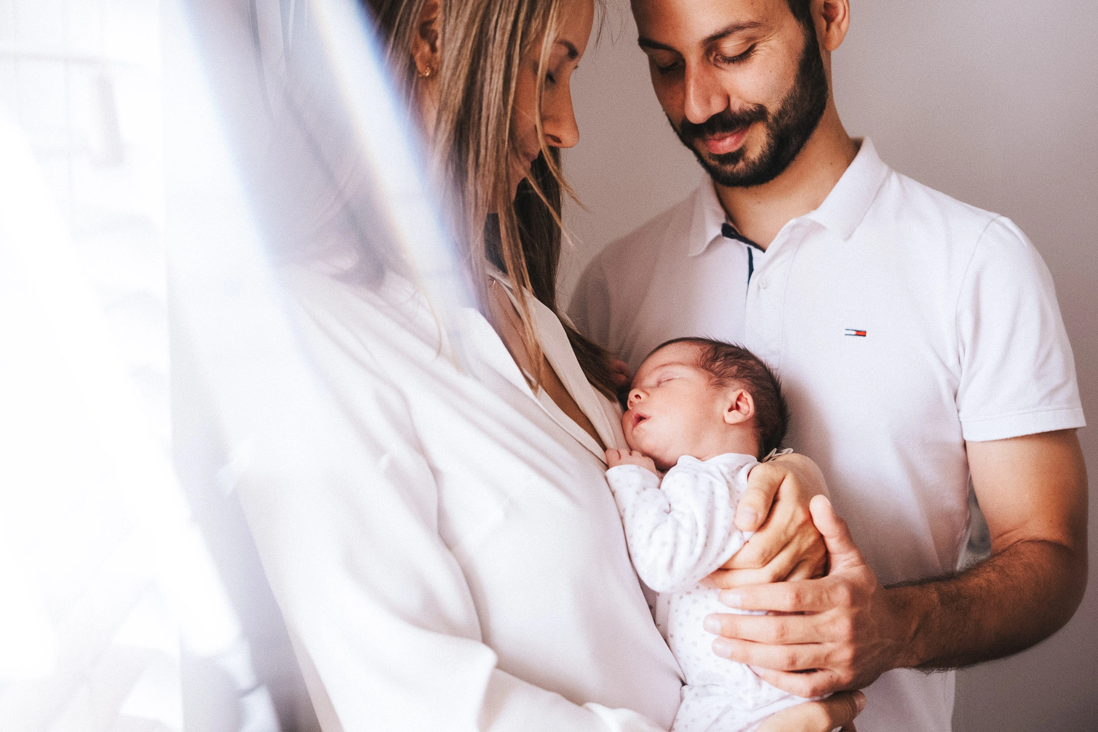 Deux parents regardent leur nouveau-né endormi dans leurs bras près d'une fenêtre.