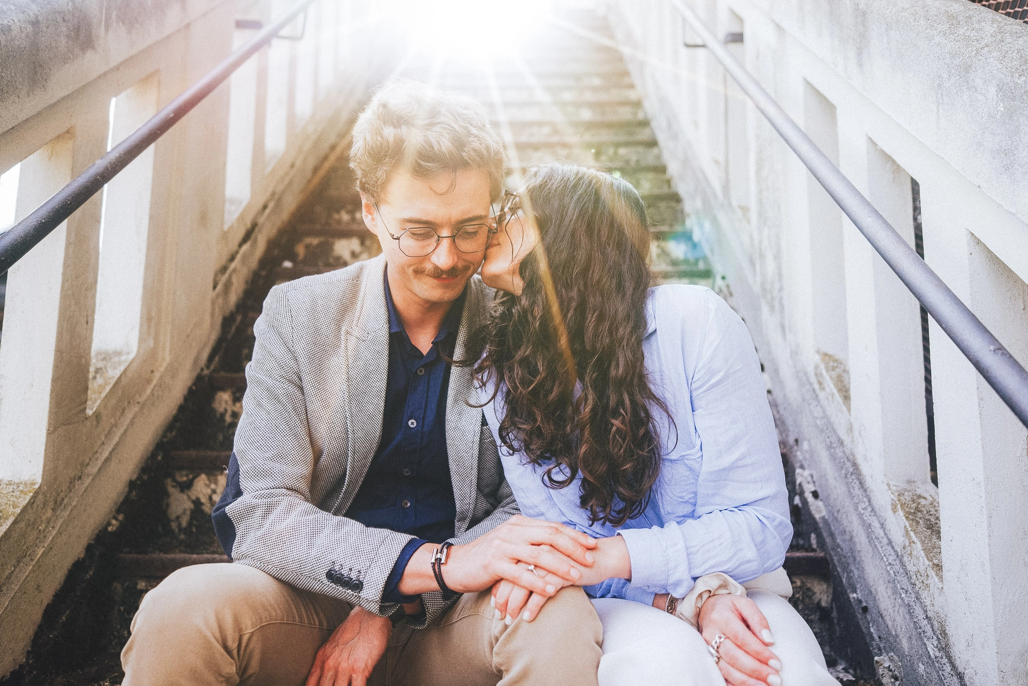 Couple assis sur un escalier extérieur, la femme embrassant son compagnon sur la joue.