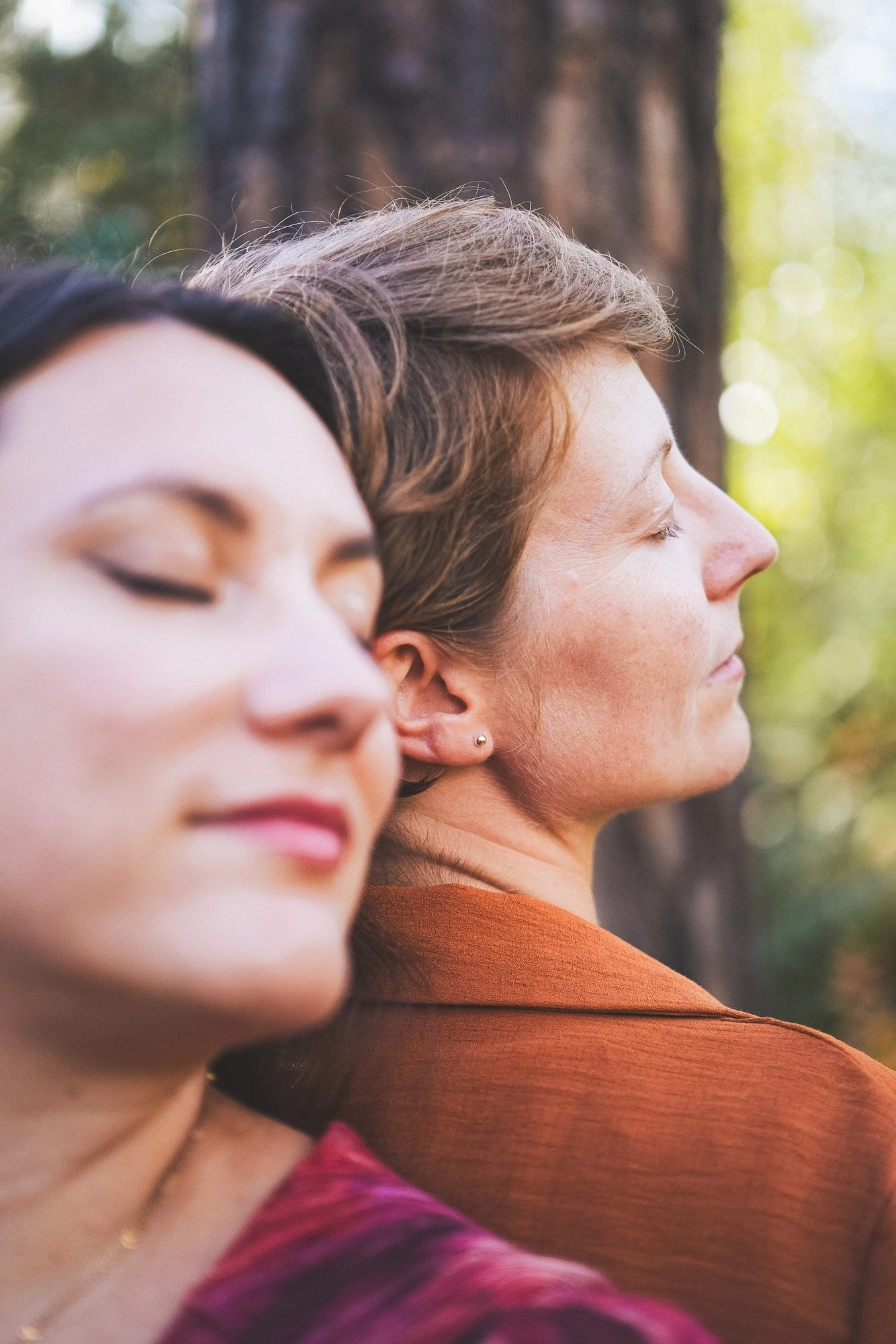 Portrait rapproché de deux femmes les yeux fermés devant des arbres baignés de lumière.