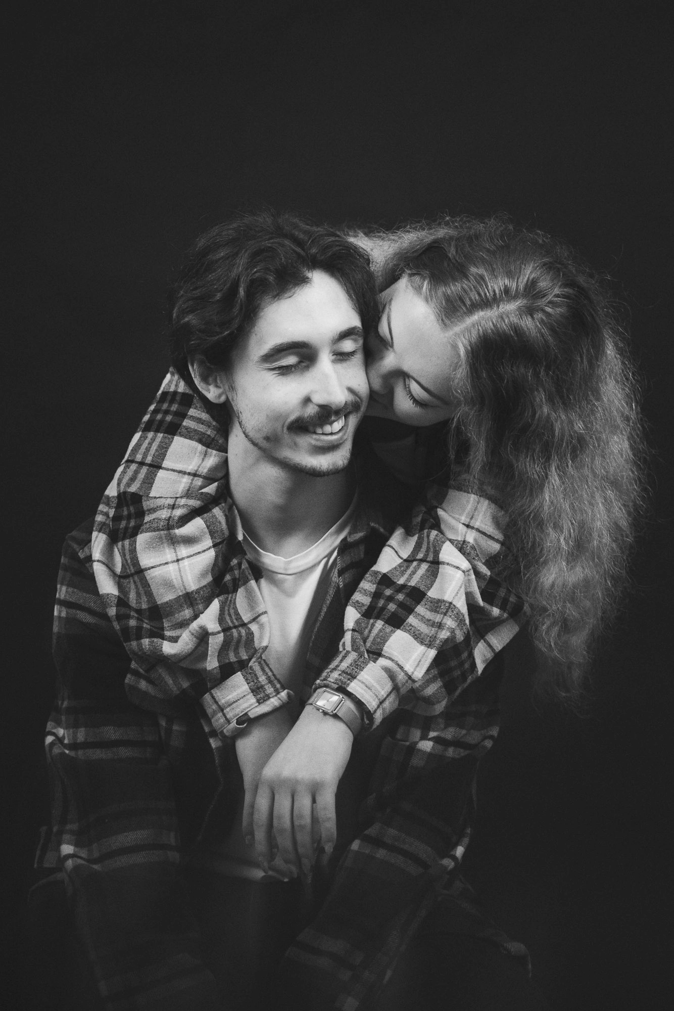 Portrait en noir et blanc d’une femme en chemise à carreaux embrassant la joue de son compagnon souriant.