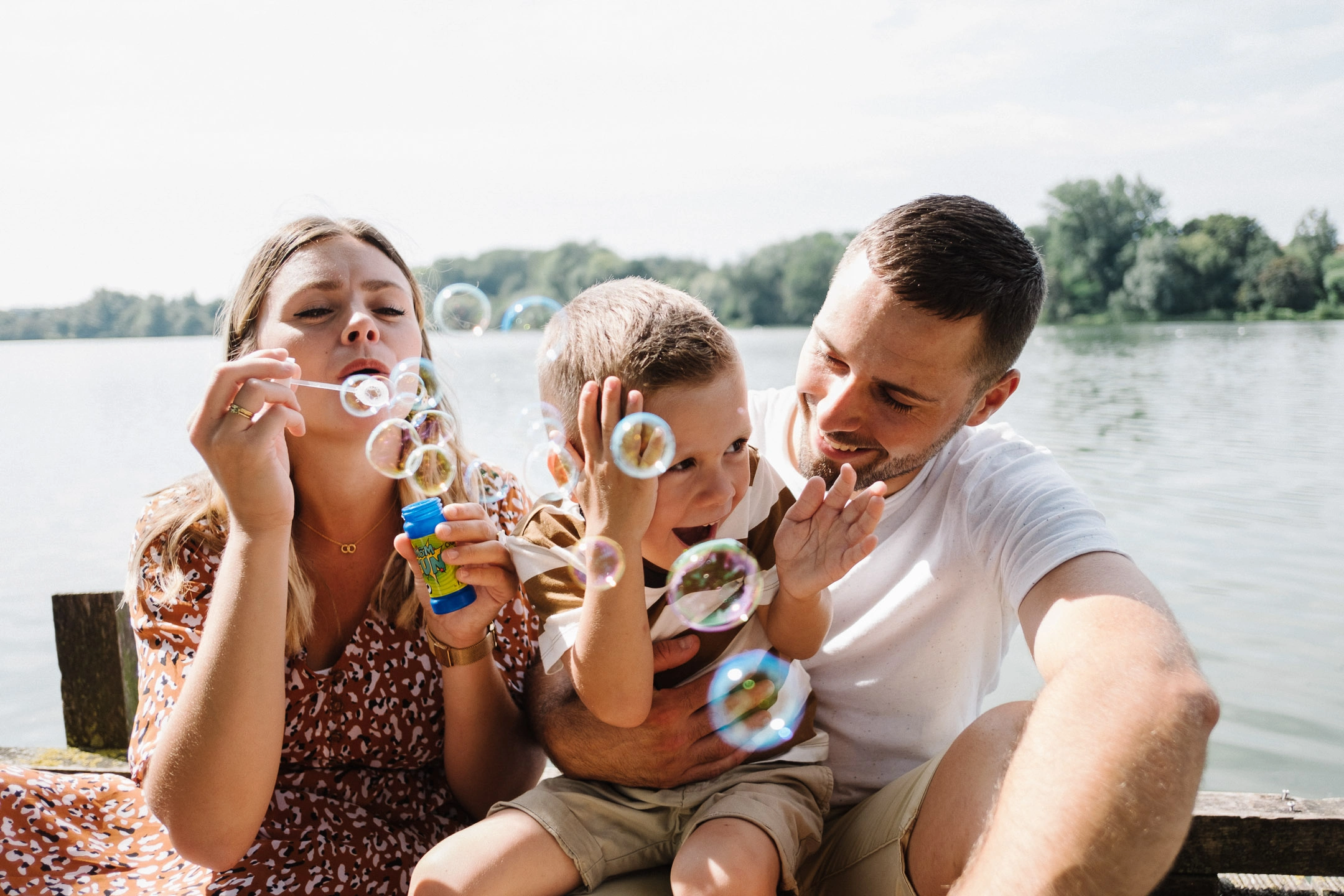 Deux parents et leur jeune garçon jouant avec des bulles de savon au bord d'un plan d'eau.