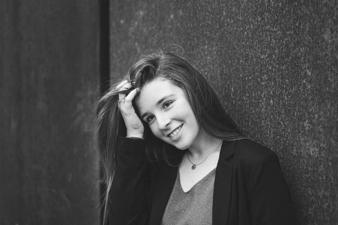 Portrait en noir et blanc d’une femme souriante appuyée contre un mur sombre.