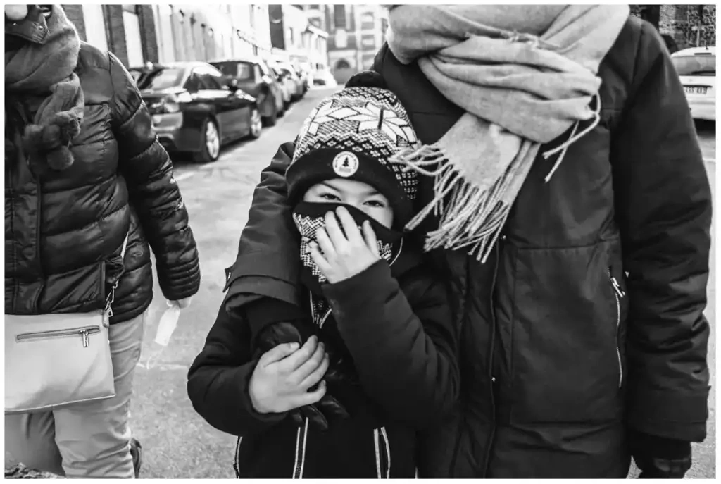 Un enfant emmitouflé dans un bonnet et une écharpe regarde l’appareil dans une rue, en noir et blanc.