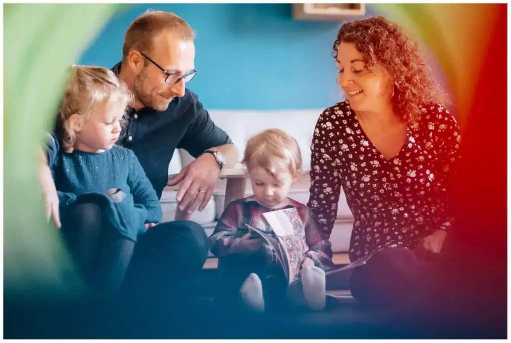 Deux parents assis avec leurs deux jeunes filles qui regardent un livre dans un salon coloré.
