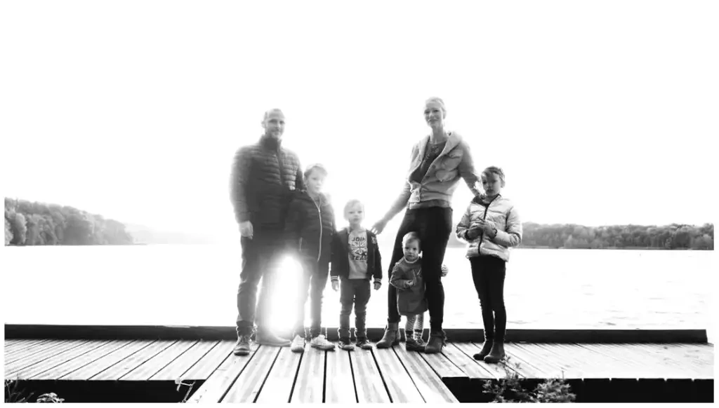 Deux adultes et quatre enfants se tiennent sur un ponton devant un étang, en noir et blanc.