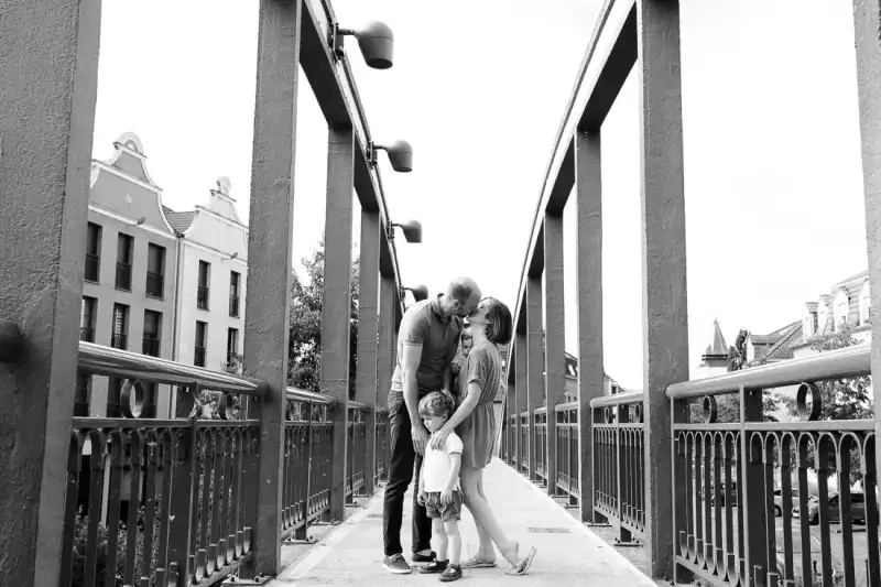 Deux parents et leur jeune enfant réunis au centre d'une passerelle, en noir et blanc.
