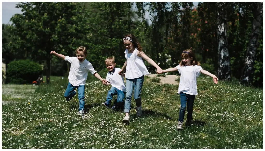Quatre enfants courent main dans la main dans une prairie parsemée de fleurs blanches.