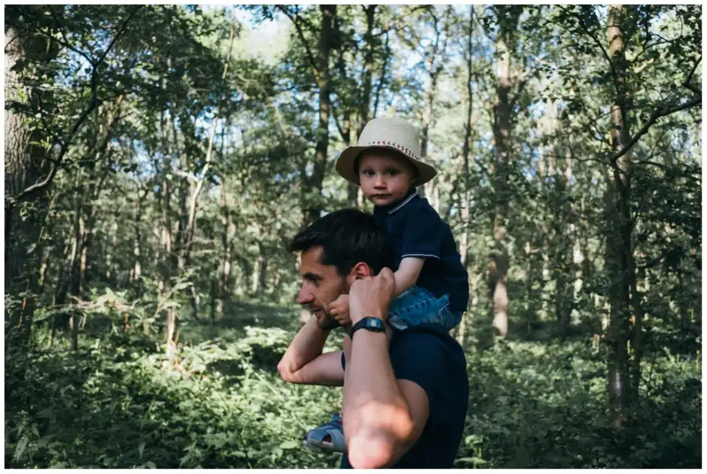 Un homme porte un jeune garçon coiffé d’un chapeau sur ses épaules dans un sous-bois.