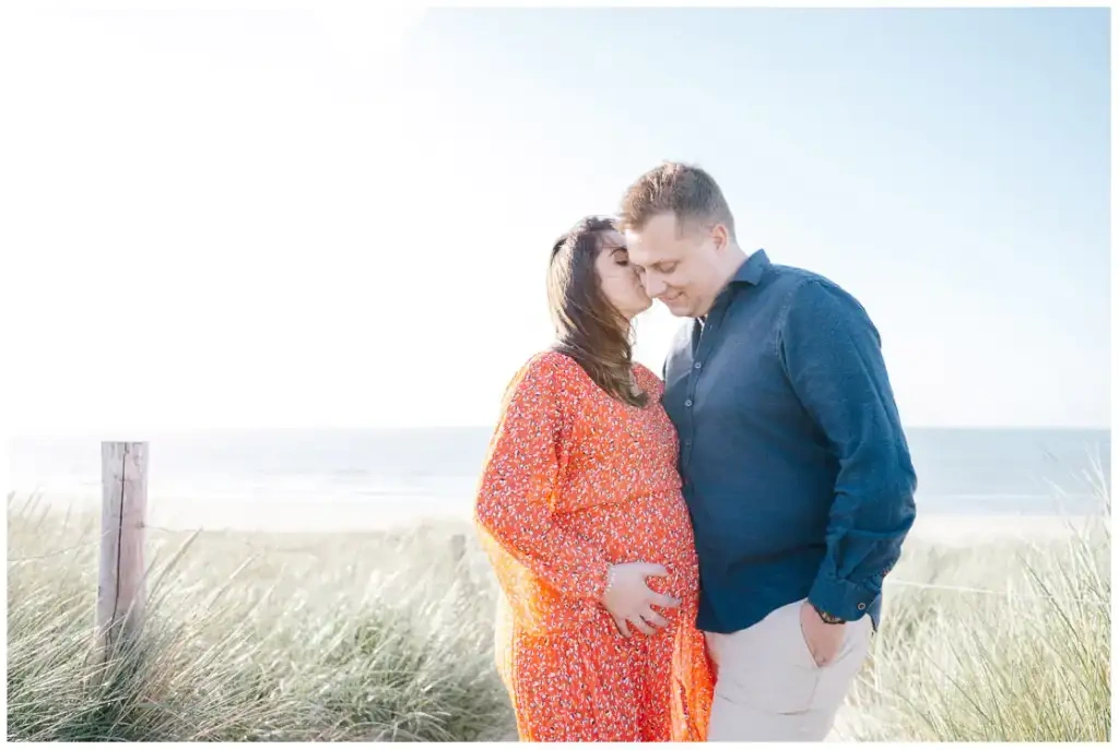Une femme enceinte en robe rouge embrasse son compagnon dans les dunes face à la mer.