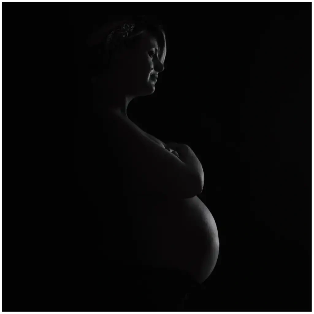 Silhouette de profil d'une femme enceinte éclairée par un fin rai de lumière sur fond noir.