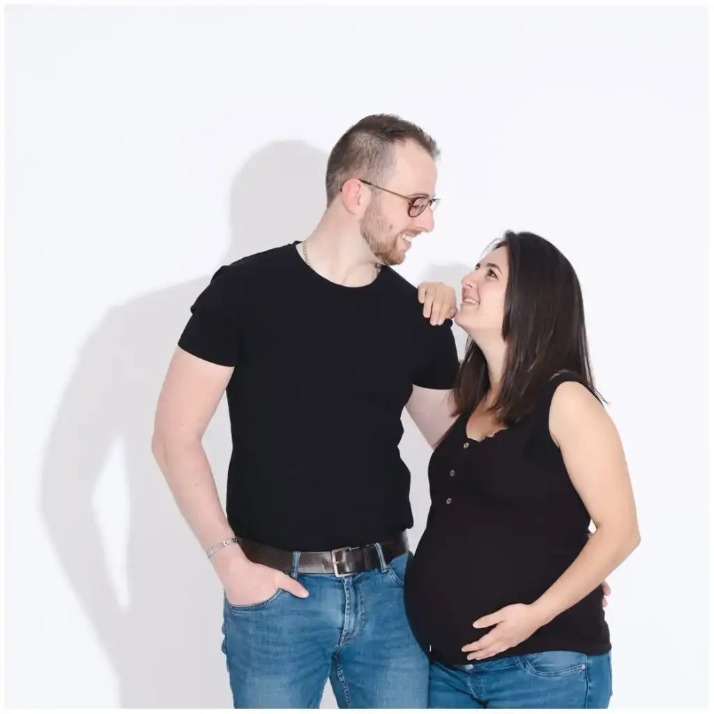 Un couple souriant se regarde devant un fond blanc pendant une séance grossesse.