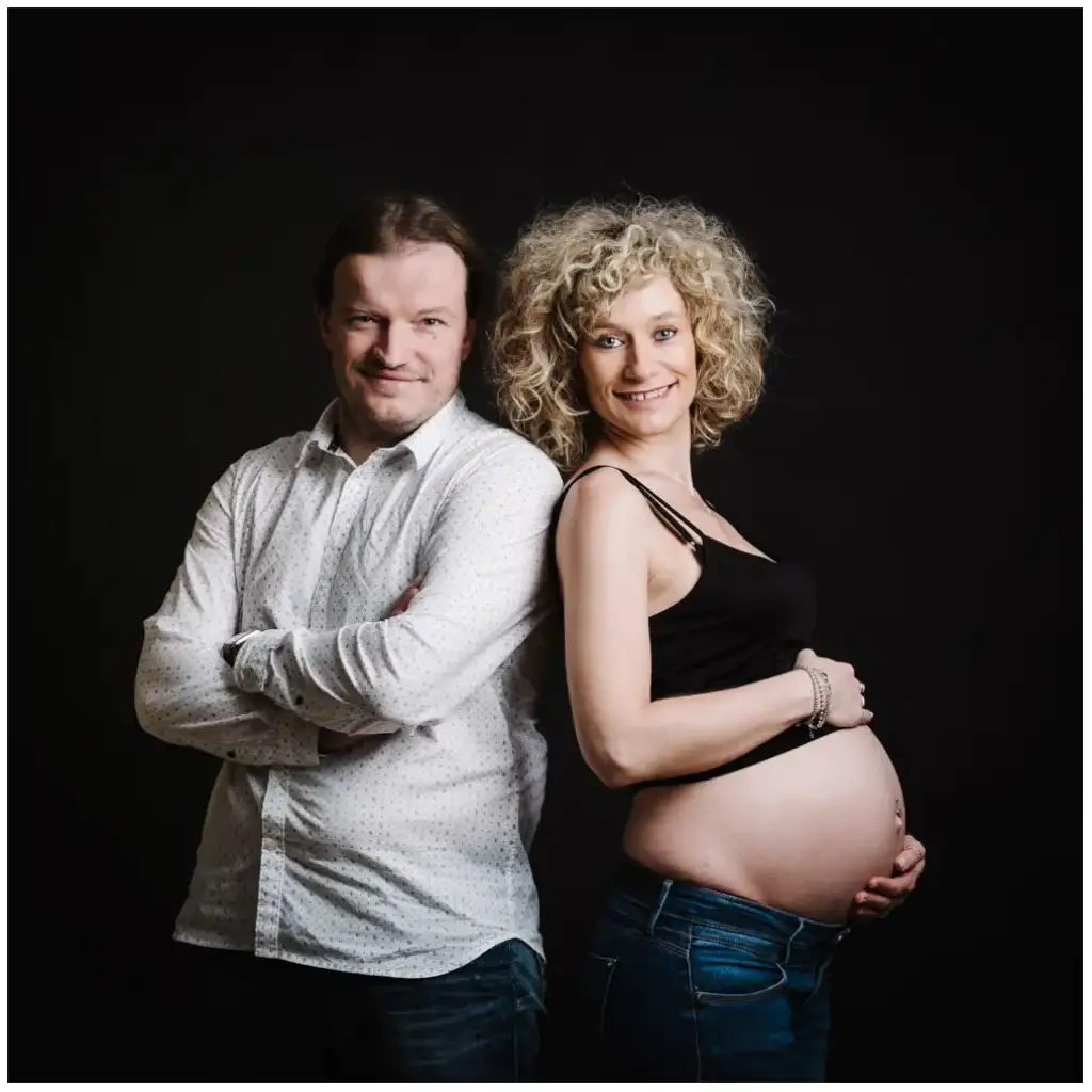Un couple se tient dos à dos devant un fond noir, la future mère posant une main sur son ventre.