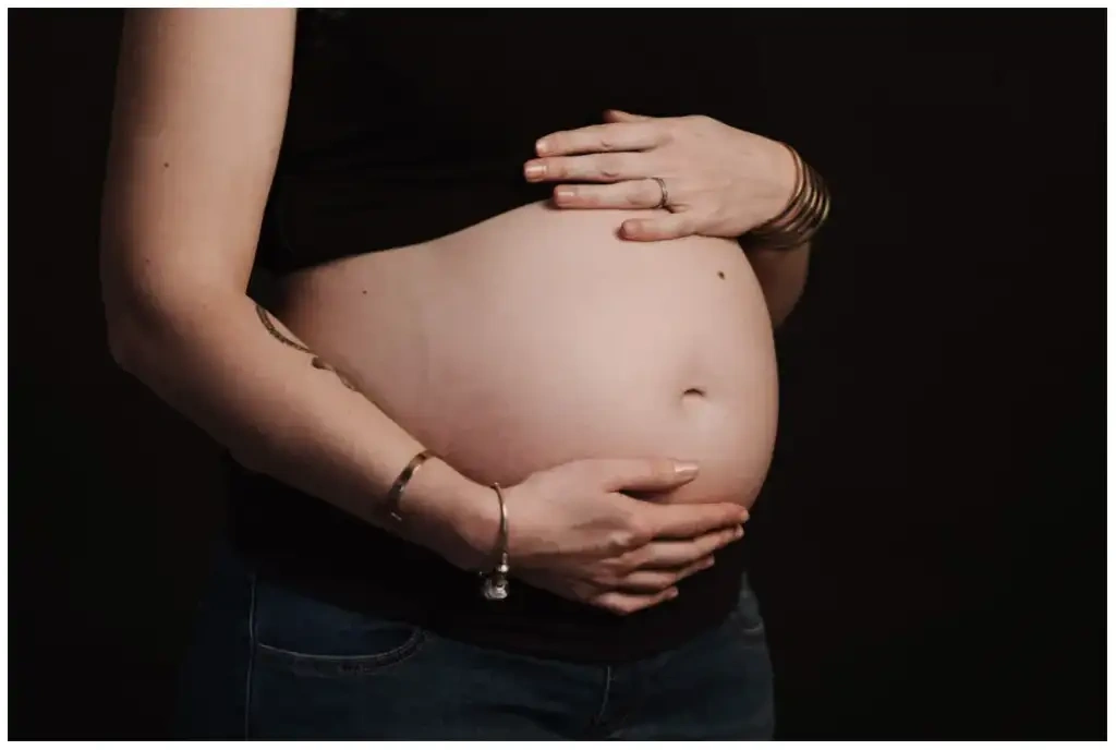 Gros plan sur le ventre d'une femme enceinte entouré de ses mains devant un fond sombre.