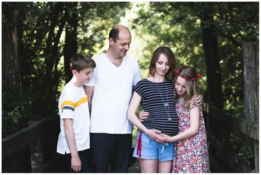 Deux parents et leurs deux enfants réunis autour du ventre de grossesse sur un sentier boisé.