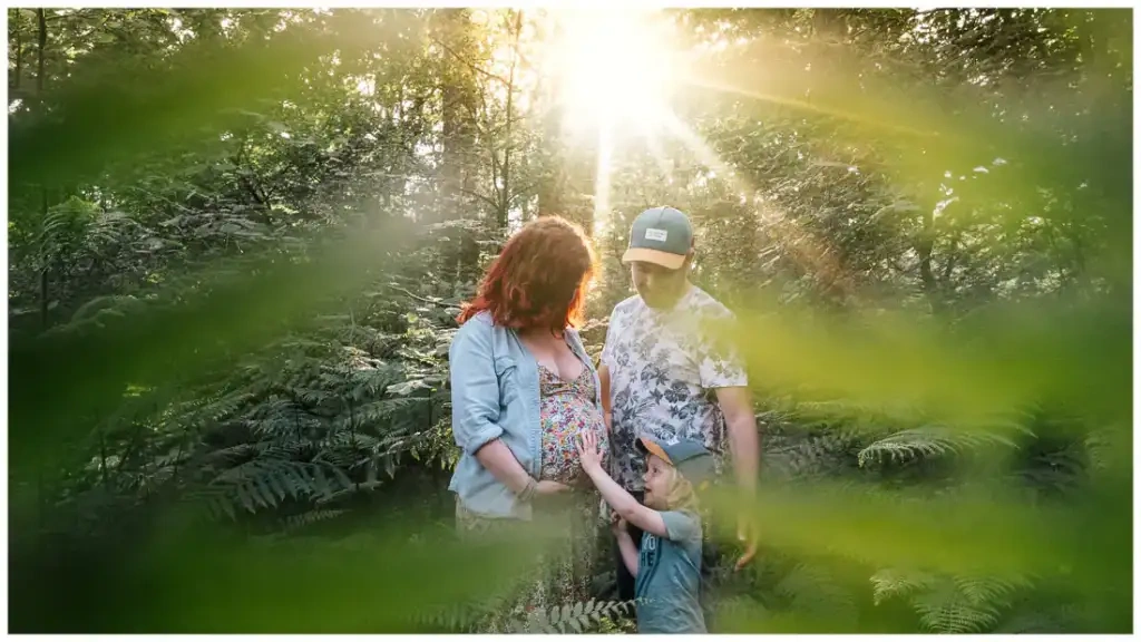 Un couple et son jeune garçon entourent le ventre de grossesse dans une forêt éclairée par le soleil.
