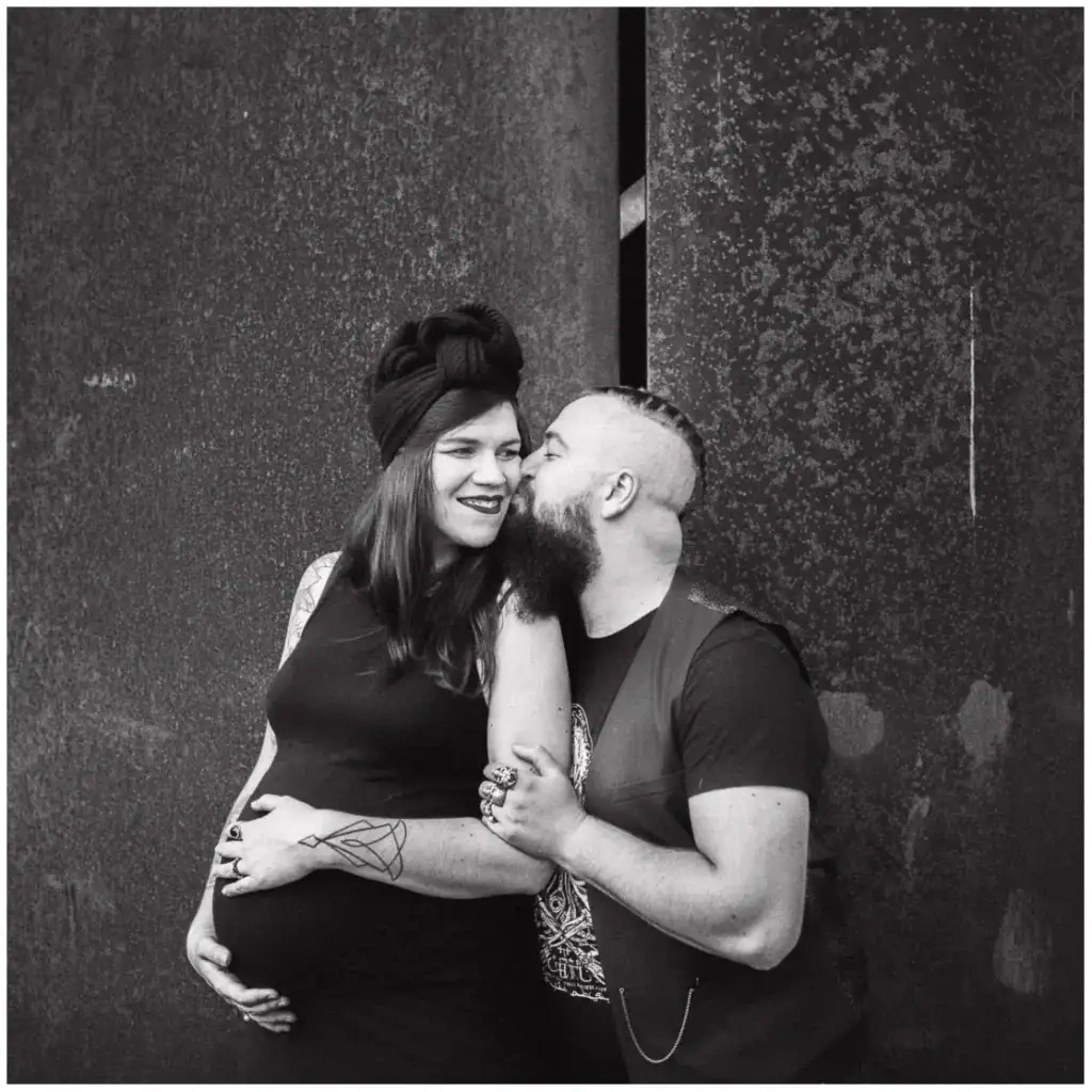 Un homme embrasse la joue de sa compagne enceinte devant une paroi métallique, dans un portrait en noir et blanc.
