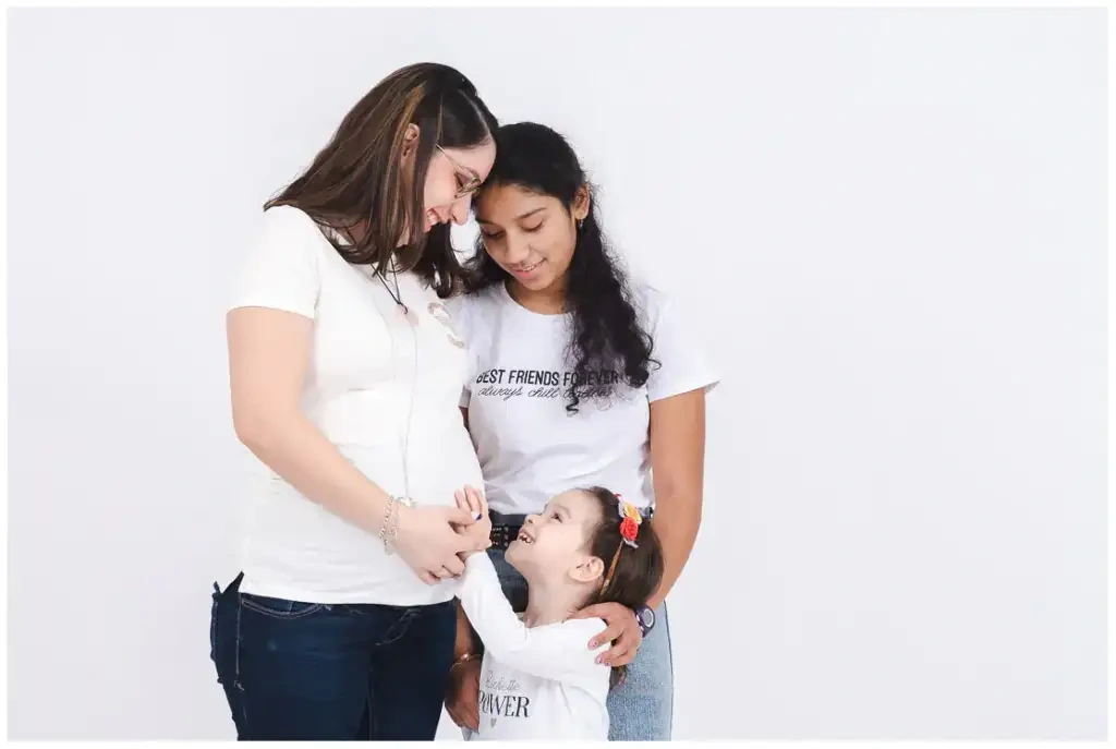 Une femme enceinte en blanc accompagnée d'une amie et d'une jeune fille devant un fond clair.