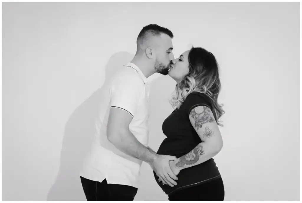Portrait en noir et blanc d'un couple qui s'embrasse, les mains réunies sur le ventre de grossesse.