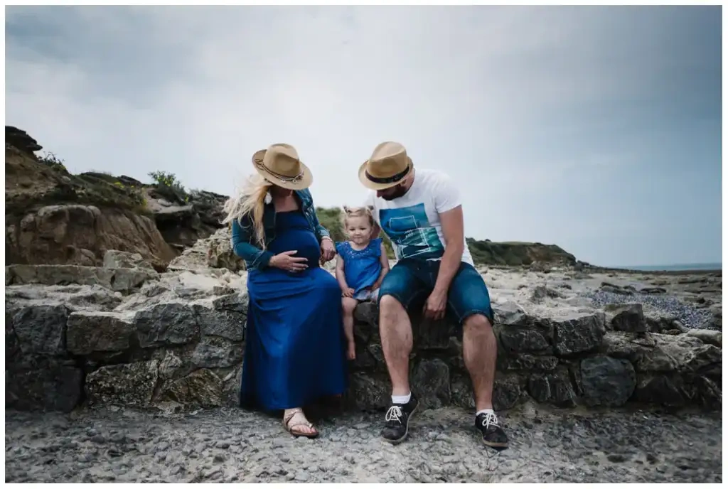 Deux parents et leur petite fille assis sur un muret rocheux au bord de la mer.