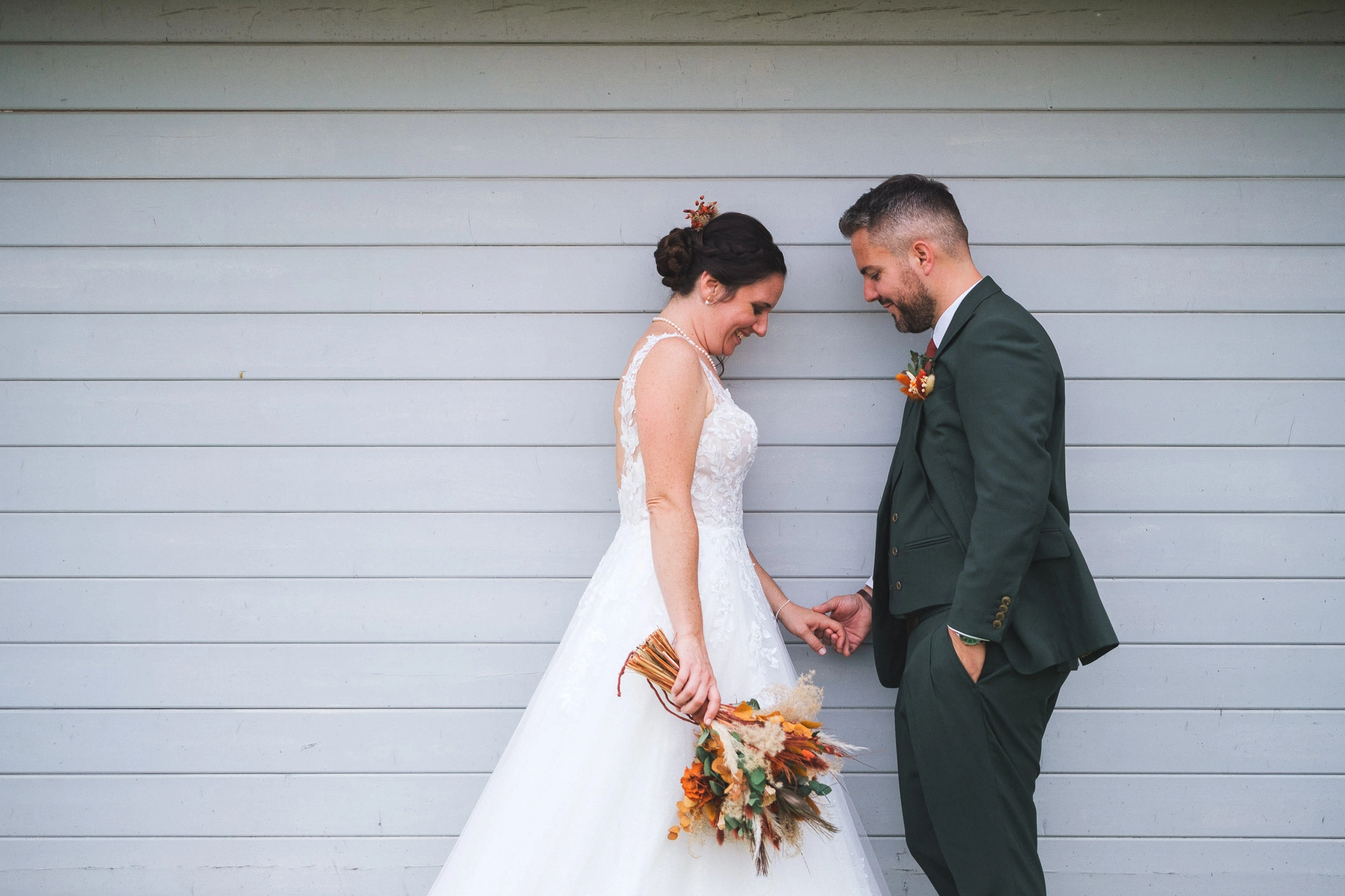 Camille et Florian se tiennent la main devant un mur en bois gris, bouquet orangé à la main.