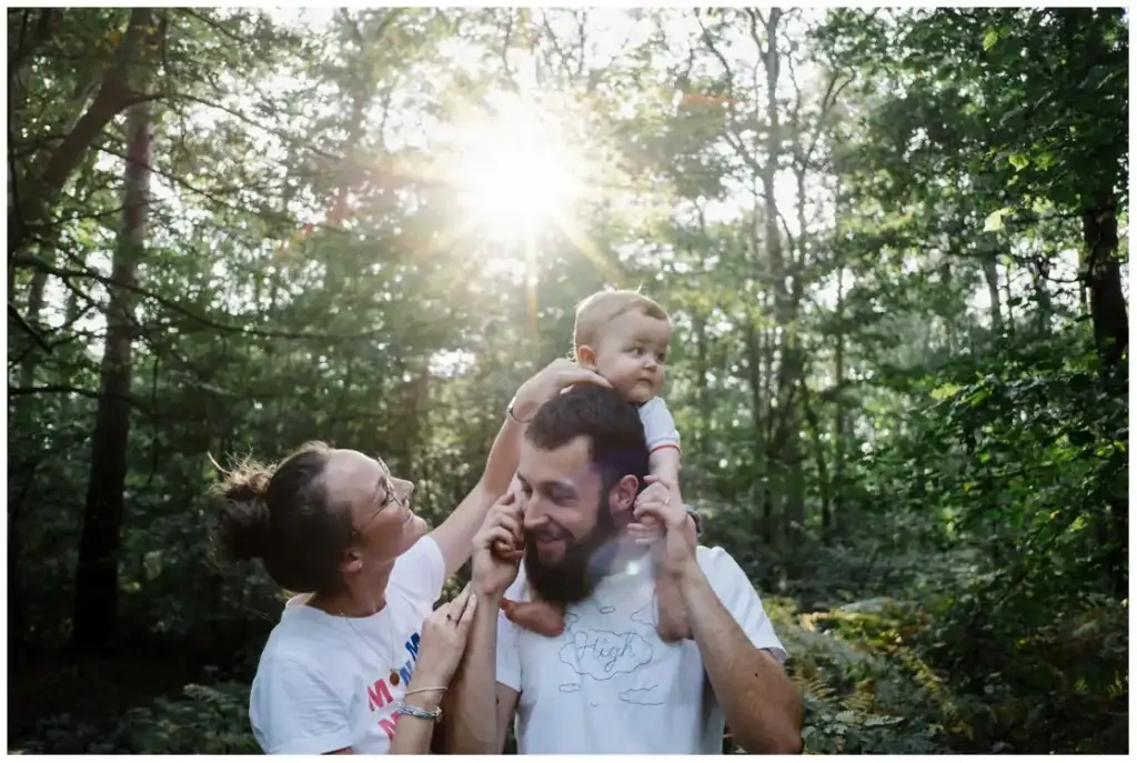 Deux parents entourent leur bébé assis sur les épaules de son père dans une forêt.