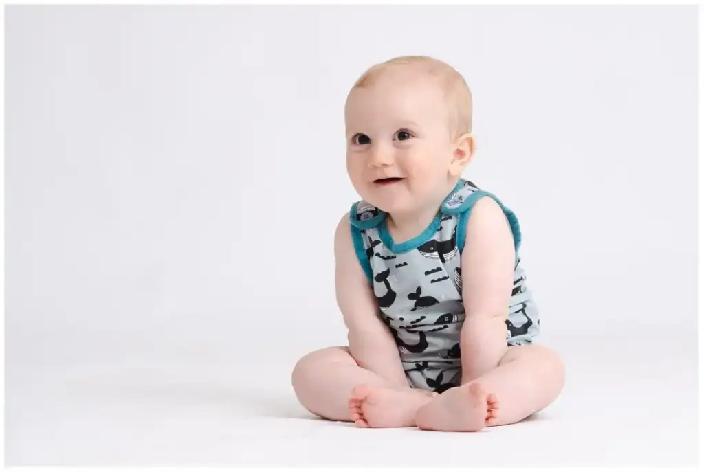 Un bébé souriant est assis en tailleur devant un fond blanc.