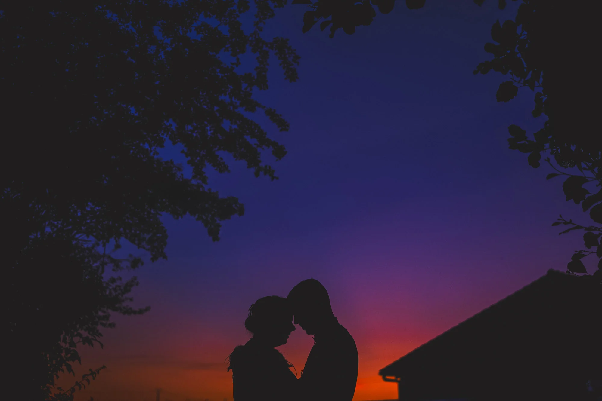 Silhouette de jeunes mariés front contre front sous un ciel bleu et orangé au crépuscule.